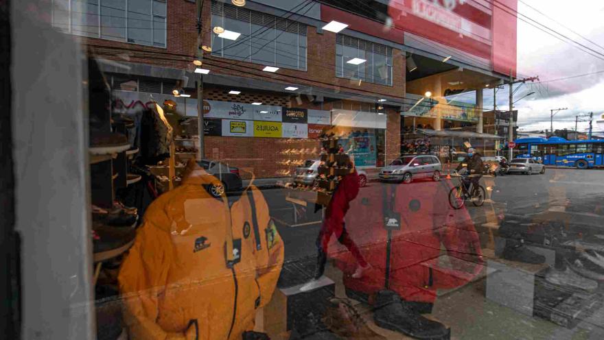 El escaparate de una tienda deportiva en Floresta Outlet exhibe prendas y calzado para actividades al aire libre, mientras el reflejo del exterior permite apreciar el movimiento de la calle, los vehículos y el transporte público frente al establecimiento.