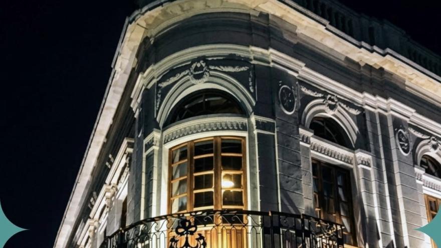 Fachada de un edificio histórico iluminado de noche, con ventanales de madera, balcones de hierro forjado y ornamentación clásica.