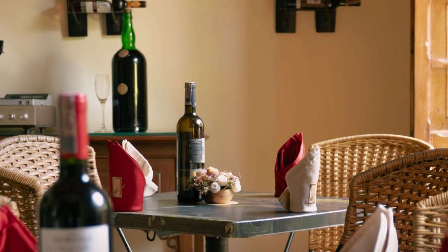 Wine bottles and glasses decorate a cozy area of the restaurant, accompanied by tables with tablecloths and floral details.