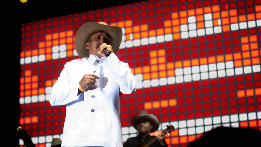 Cantante con traje blanco y sombrero interpreta con micrófono frente a una pantalla luminosa en concierto nocturno.