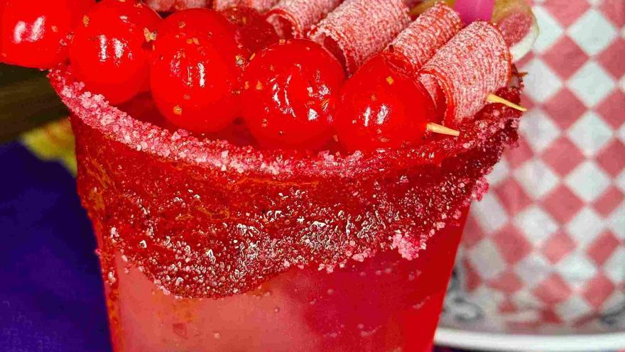 Bebida roja decorada con frutas, dulces y un borde cubierto de chile.