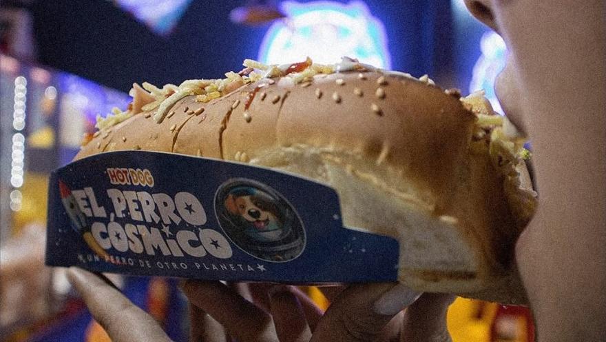 Perro caliente de El Perro Cósmico servido con pan de ajonjolí y abundantes toppings.