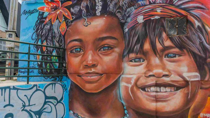 Mural de arte urbano con los rostros de dos niños, rodeados de flores, colores y elementos de la cultura local.
