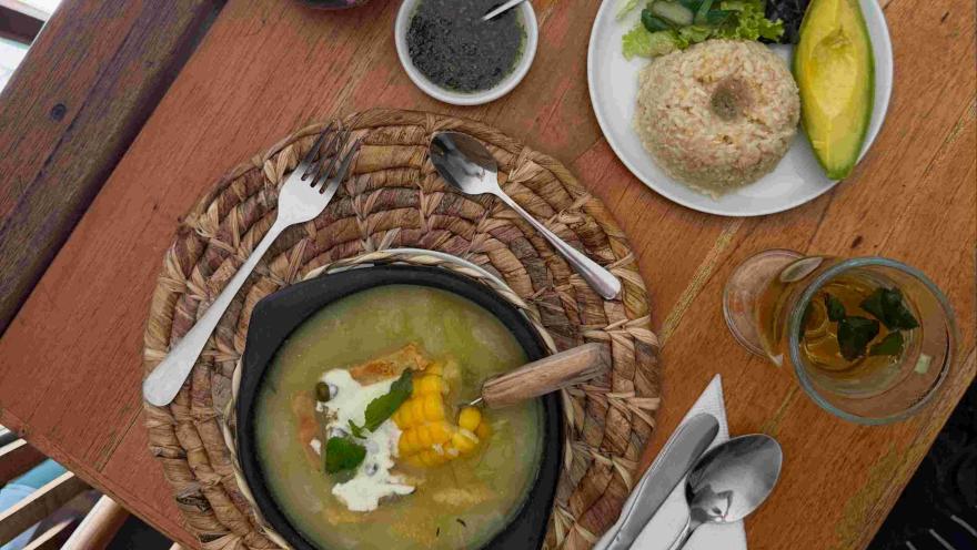  Plato vegetariano como ajiaco con sopa de vegetales, maíz, crema y hierbas, acompañado de ensalada, aguacate y una bebida caliente.