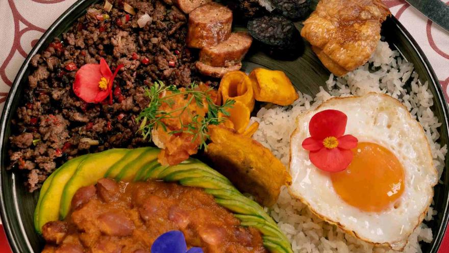 Traditional Colombian dish with rice, beans, ground beef, fried egg, avocado, chorizo, plantain, and pork belly, garnished with edible flowers.