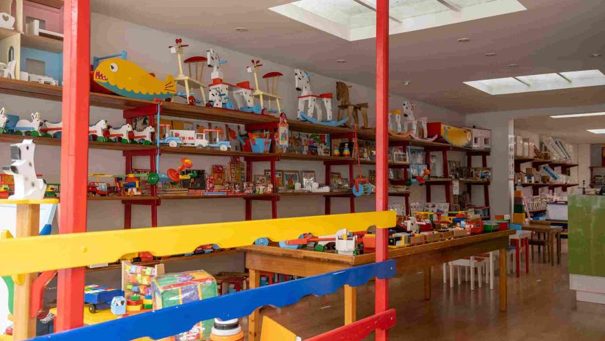 La imagen muestra el interior de una colorida juguetería, con estanterías repletas de juguetes tradicionales, juegos didácticos y figuras de madera. El espacio, iluminado por tragaluces y organizado alrededor de mesas y exhibidores, transmite un ambiente alegre y familiar.