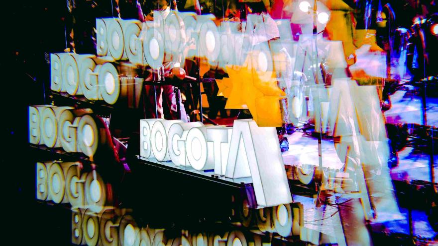 Escenario con luces y músicos en concierto, con la palabra “Bogotá” repetida en un efecto visual colorido y superpuesto.