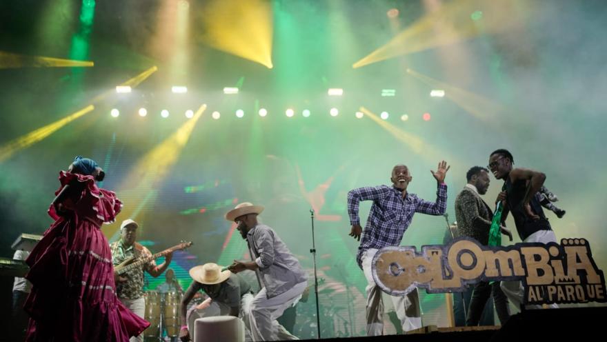 Grupo de músicos y bailarines se presenta en un escenario con luces de colores durante un concierto.