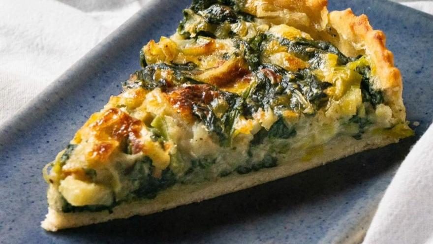 Tarta salada de masa horneada, cubierta con espinaca, vegetales y queso, presentada sobre un plato azul.