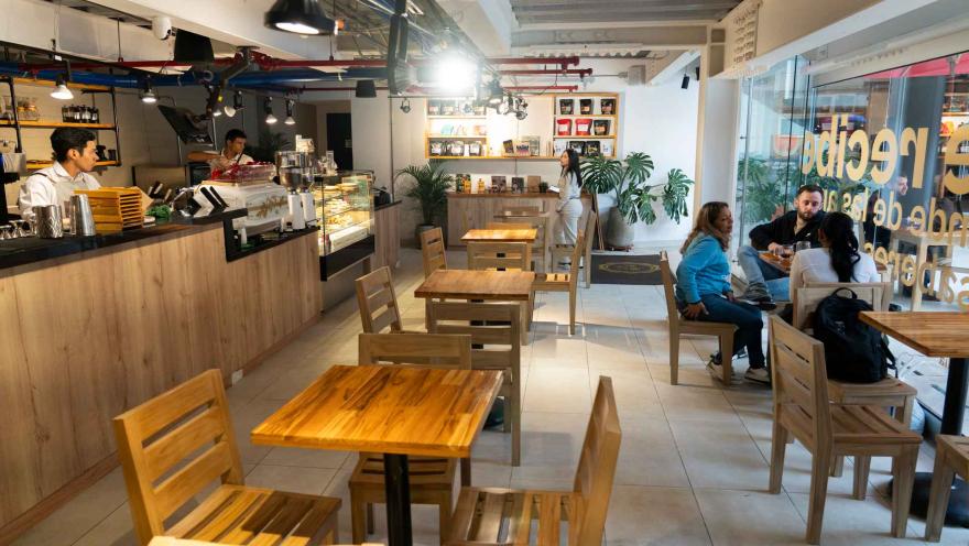 Salón interior iluminado con mesas de madera, mostrador al fondo, estantes de café en bolsa y clientes en la entrada.