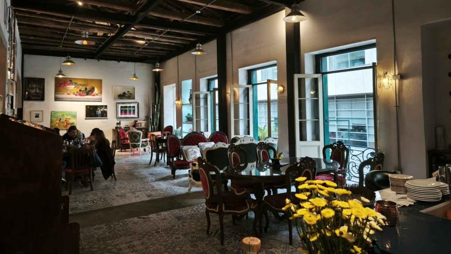 Salón de época con sillas de estilo clásico, grandes ventanales, pinturas en las paredes y flores amarillas. 