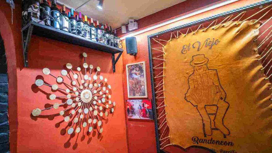Pared roja decorada con adornos metálicos, botellas de vino en estante y tapiz de cuero con el logo de El Viejo.
