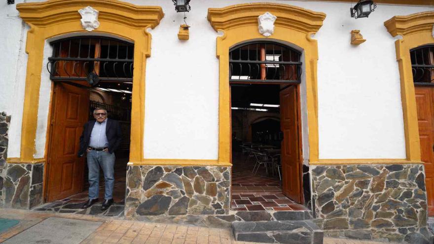 Hombre mayor con gafas y saco oscuro parado en la puerta de entrada de una casona colonial con detalles amarillos.