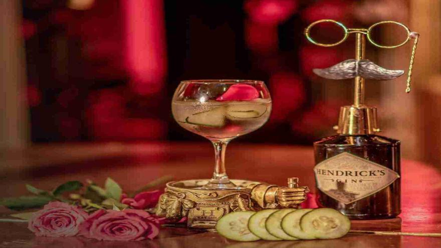 Un elegante cóctel de ginebra con pepino y pétalos de rosa, acompañado de una botella de Hendrick's Gin y adornos vintage, sobre un fondo rojo romántico.