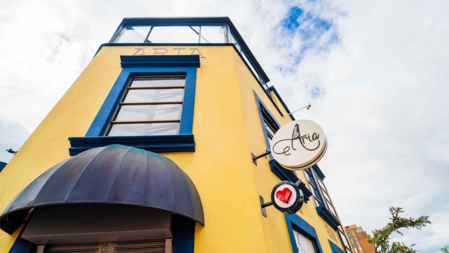 Fachada de un edificio amarillo de esquina con marcos de ventanas azules, marquesina oscura y letreros circulares de la marca Aria.