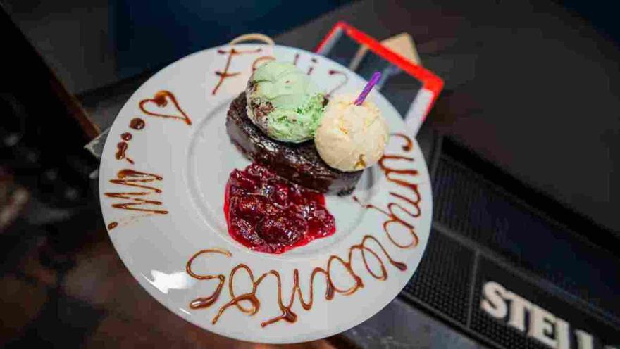 Postre de brownie con dos bolas de helado, salsa de frutos rojos y mensaje de Feliz Cumpleaños escrito en caramelo.