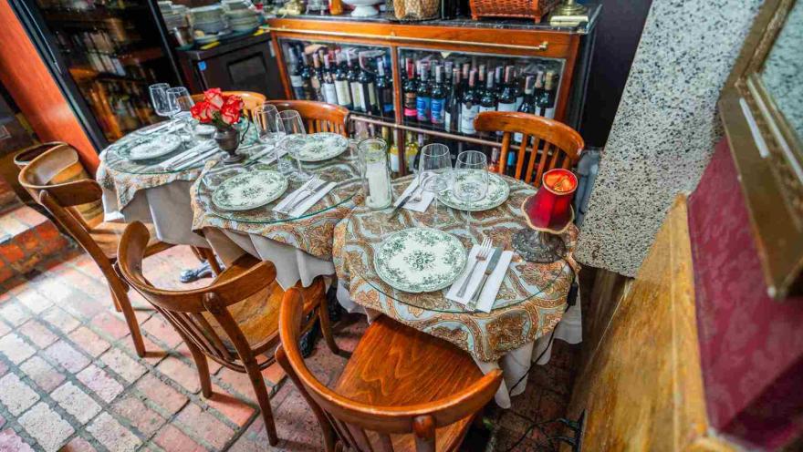 Mesas redondas de madera servidas con manteles estampados, vajilla decorativa y copas de cristal en un salón rústico.