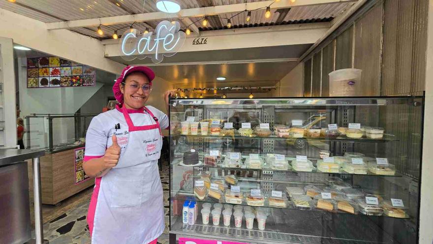  Repostera con gorro y delantal fucsia sonríe haciendo un gesto de aprobación al lado de una vitrina de postres. 