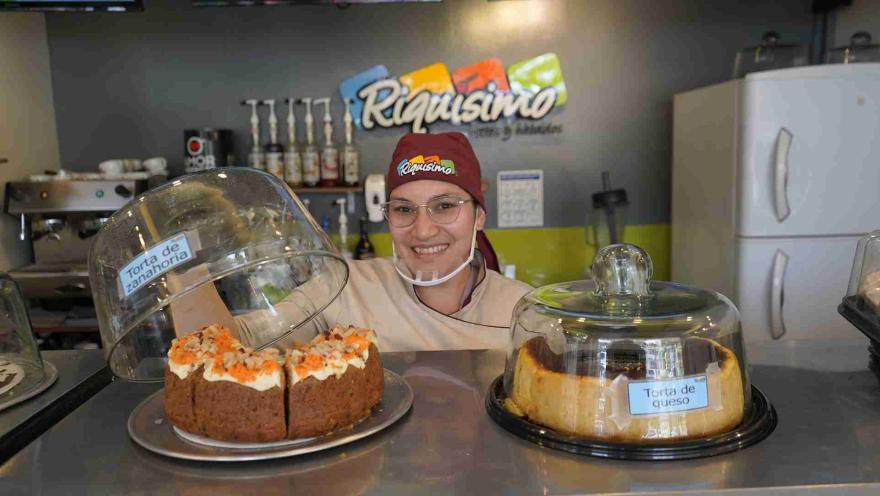 Pastelera sonriente con uniforme detrás del mostrador sosteniendo una campana de vidrio sobre una torta de zanahoria. 