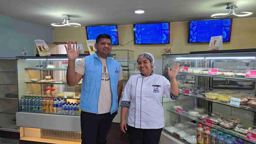  Un hombre con chaleco azul y una mujer con uniforme de chef sonríen y saludan frente a vitrinas llenas de postres en un establecimiento llamado "La Torre de los Postres".