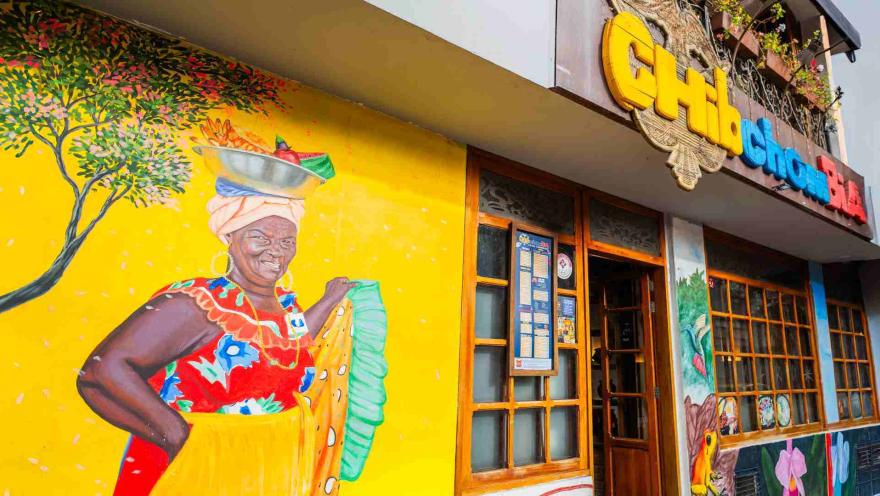 Plano medio del mural de la fachada con una mujer palenquera sonriendo sobre un fondo amarillo vibrante.