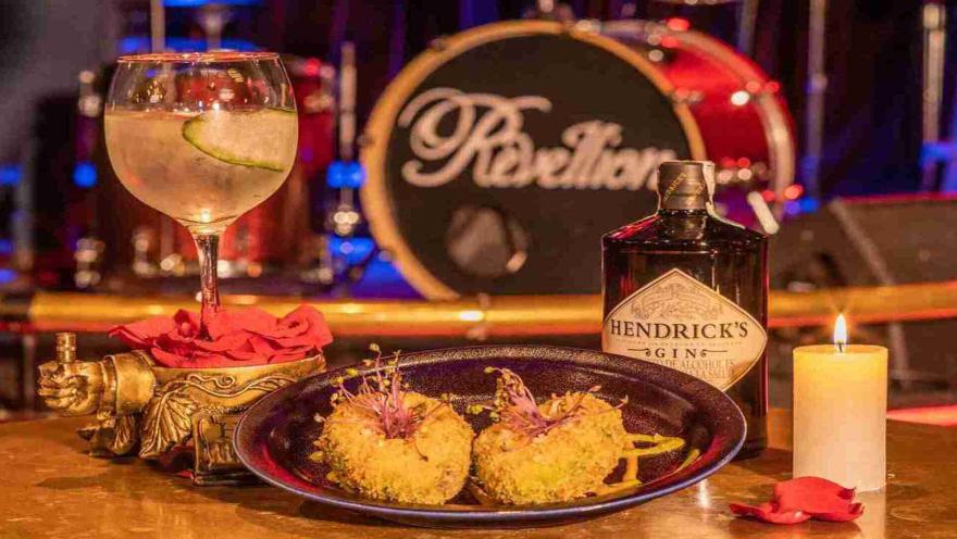 Un cóctel de ginebra y Hendrick's, junto a dos aperitivos empanizados, una vela y pétalos de rosa, con una batería de fondo que dice "Rebellion".