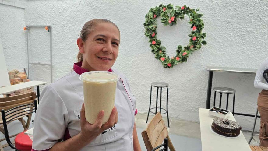Mujer con filipina de cocina sostiene un vaso transparente con malteada frente a una pared con corazón floral. 