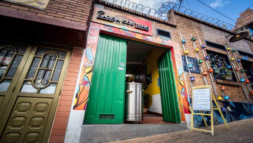  Vista en ángulo de la entrada del restaurante Sazón Costeño con letrero, marco decorado y tablero de menú.