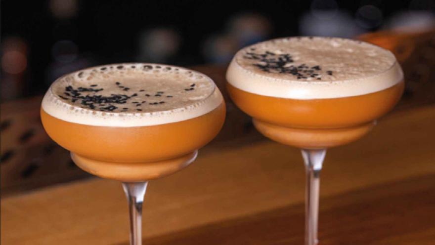 Dos copas de cóctel de tono ámbar con espuma y semillas de ajonjolí negro decorando la parte superior. 