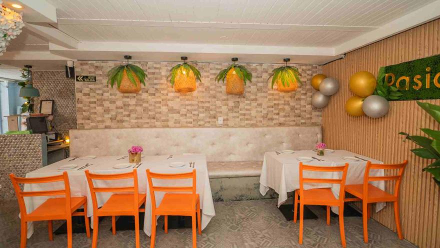 Comedor con sillas de plástico naranja, mesas con manteles blancos, pared de piedra y lámparas con follaje vegetal.