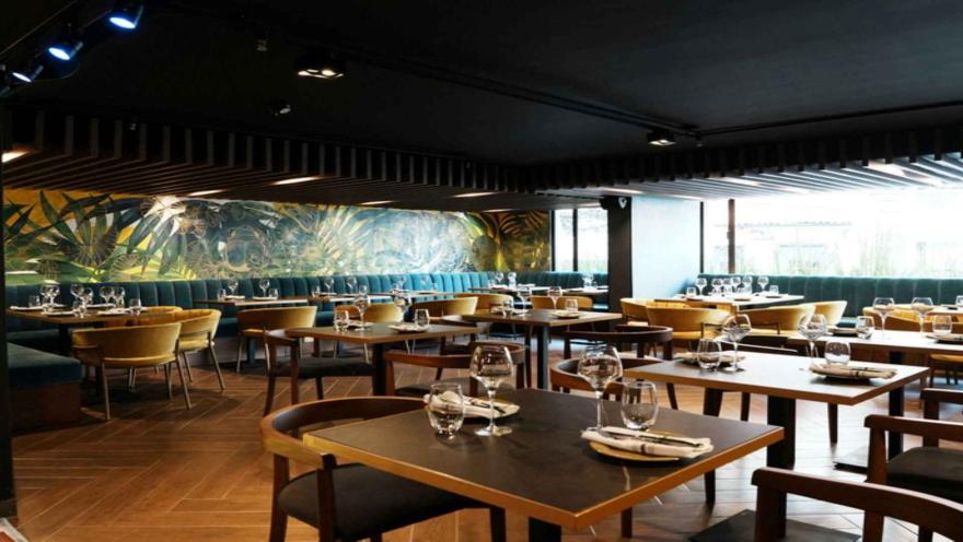 Restaurante contemporáneo con mesas de madera, amplios ventanales, iluminación cálida y un mural de inspiración tropical en el fondo.