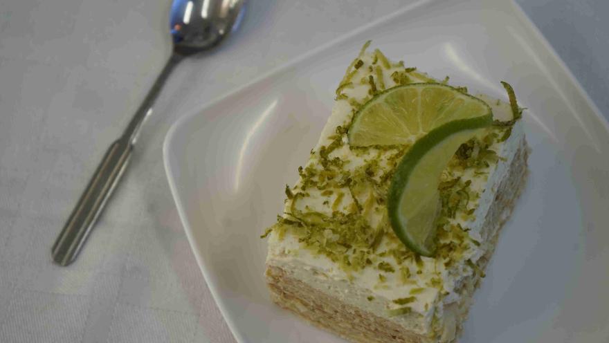 Porción cuadrada de postre de limón con ralladura y rodajas de lima sobre plato blanco. 