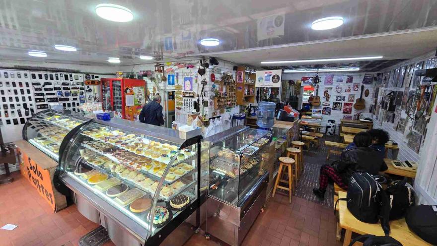 Vista panorámica del interior de la pastelería con vitrinas exhibidoras llenas de postres, barra con bancos y clientes. 