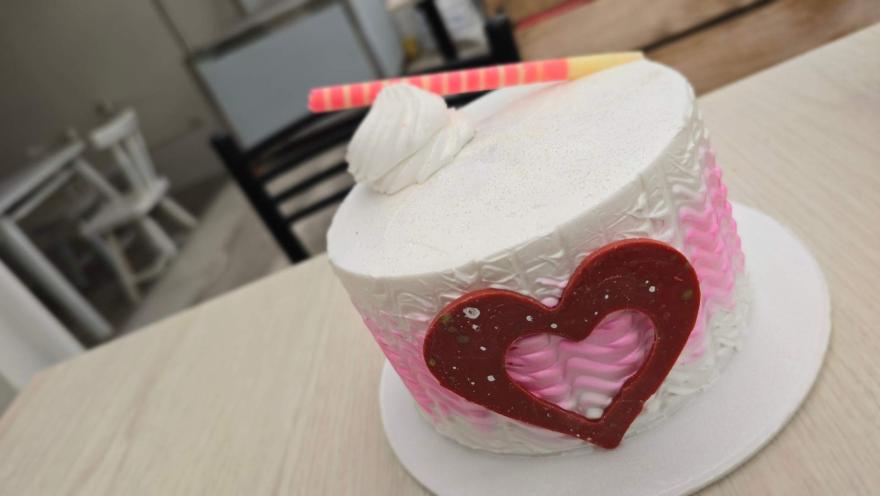Pastel cilíndrico decorado con crema blanca y rosada, detalle de corazón de chocolate rojo y espiral dulce. 