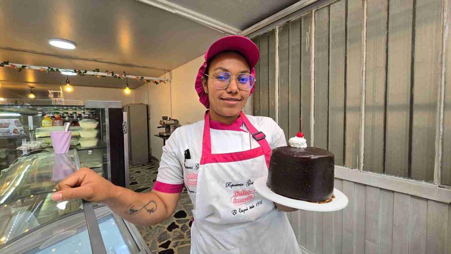 Pastelera en uniforme rosa sostiene un plato blanco con una torta de chocolate decorada con crema y una cereza. 