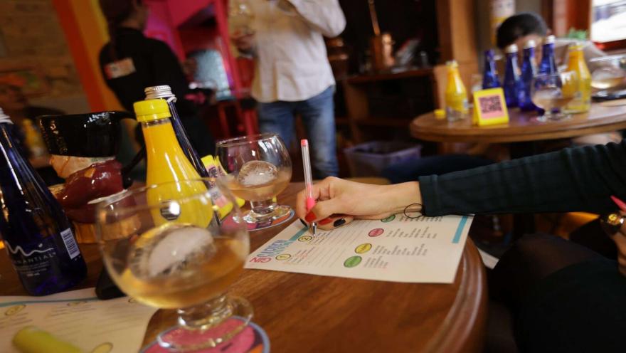 Persona tomando notas en una ficha de cata junto a copas con hielo y botellas en una mesa de madera. 