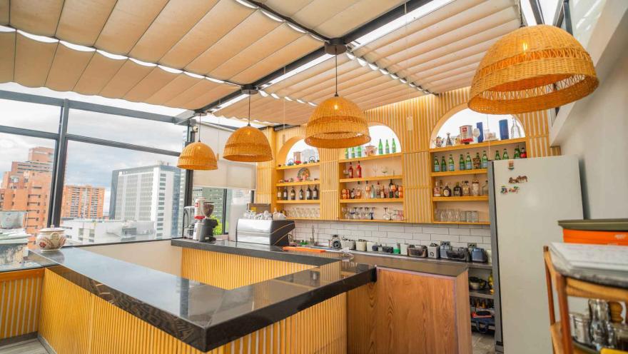 Barra de bar en la terraza superior con acabado acanalado de madera, techo retráctil y estantes iluminados con botellas.