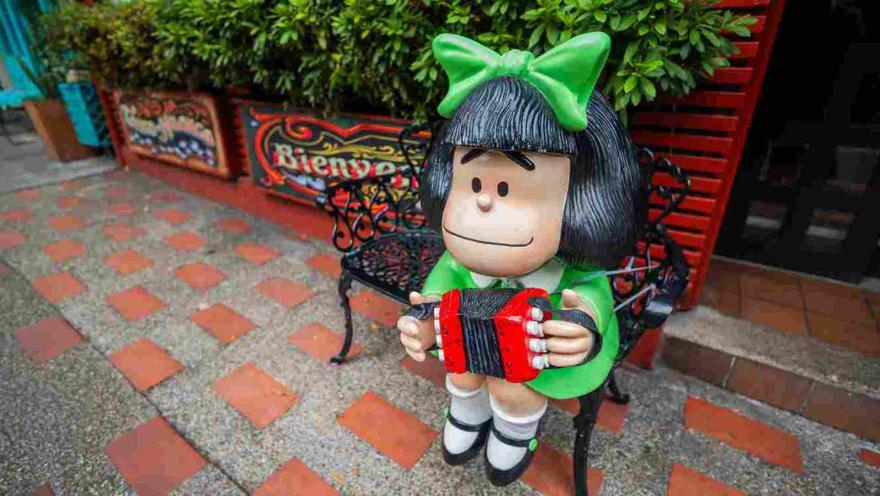 Estatua decorativa de Mafalda vestida de verde tocando un bandoneón rojo sobre una banca en la entrada.