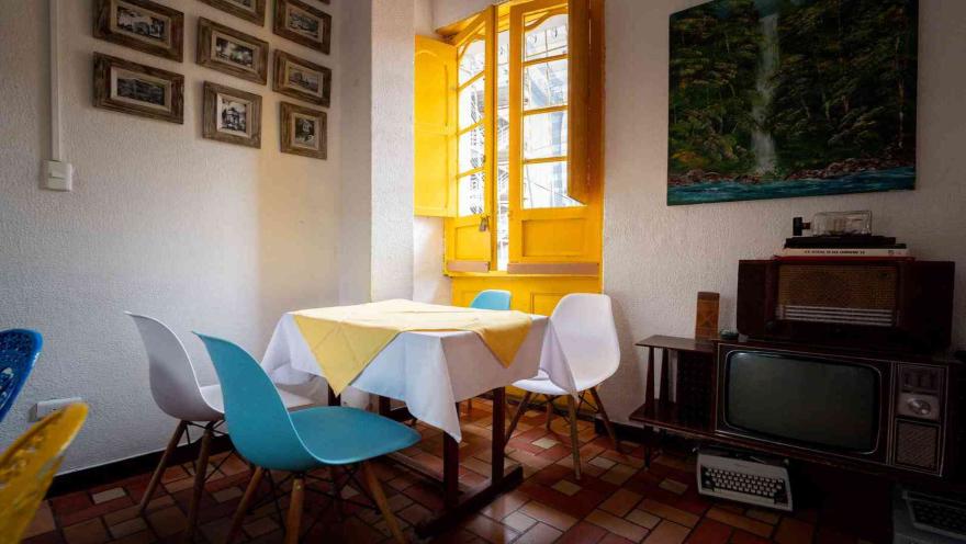  Mesa con mantel amarillo y azul junto a una ventana amarilla. Un televisor antiguo y una máquina de escribir están al lado.