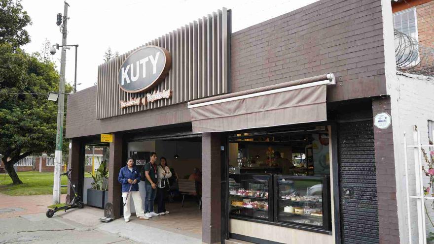  Exterior del local de Panadería Kuty con letrero iluminado, vitrina de postres y personas afuera. 