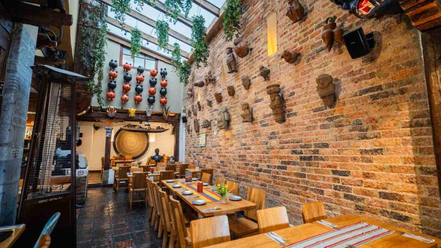 Salón amplio con iluminación natural, pared de ladrillo con esculturas, techos altos y mesas de madera ordenadas.