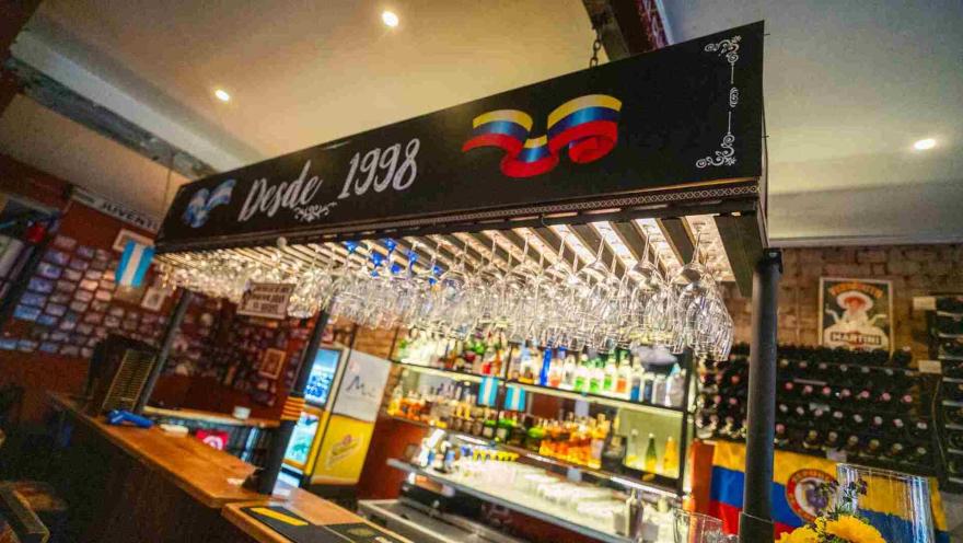 Barra del bar iluminada con copas de vino suspendidas del techo, botellas de licor y letrero con la frase "Desde 1998".