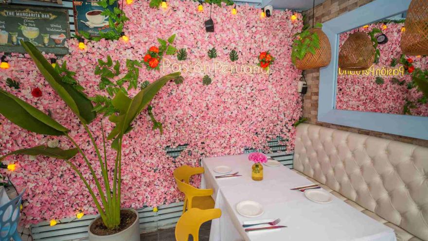 Rincón del comedor con pared cubierta de flores rosas, letrero luminoso de la marca, sillón blanco y sillas amarillas.