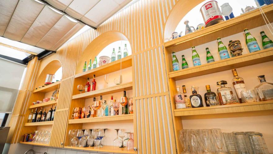  Estantería de madera iluminada con arcos, exhibiendo botellas de licor, recipientes de cobre, vasos y copas de vidrio.