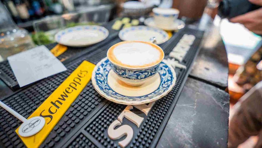  Taza de café con leche espumosa servida en porcelana decorada sobre un tapete de bar de marca Schweppes.