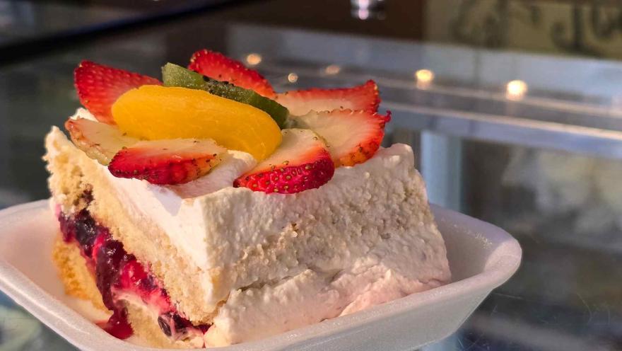 Porción de torta fría con crema, salsa de mora de fondo y rodajas de fruta fresca encima en recipiente blanco.