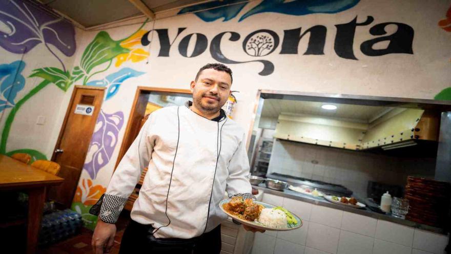 Chef con uniforme blanco sonriendo mientras sostiene un plato preparado frente al mural del restaurante Yoconta.