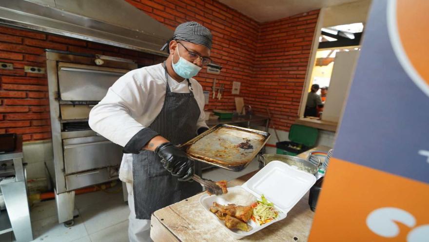 Cocinero uniformado sirviendo comida horneada desde una bandeja hacia un contenedor para llevar.