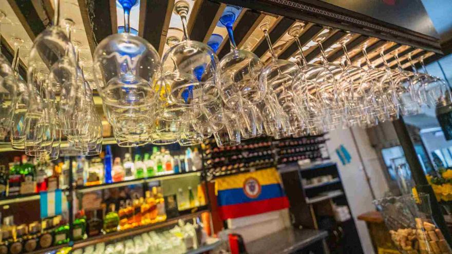 Toma en contrapicado de copas de cristal colgadas en el estante de la barra sobre botellas y banderas latinoamericanas.