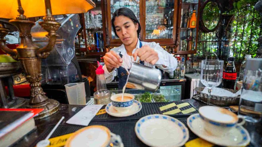 Barista vertiendo leche vaporizada en una taza de café en una barra rodeada de botellas y cristalería clásica.